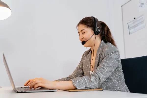 Woman in a plaid blazer working at a laptop with a headset in an office setting.