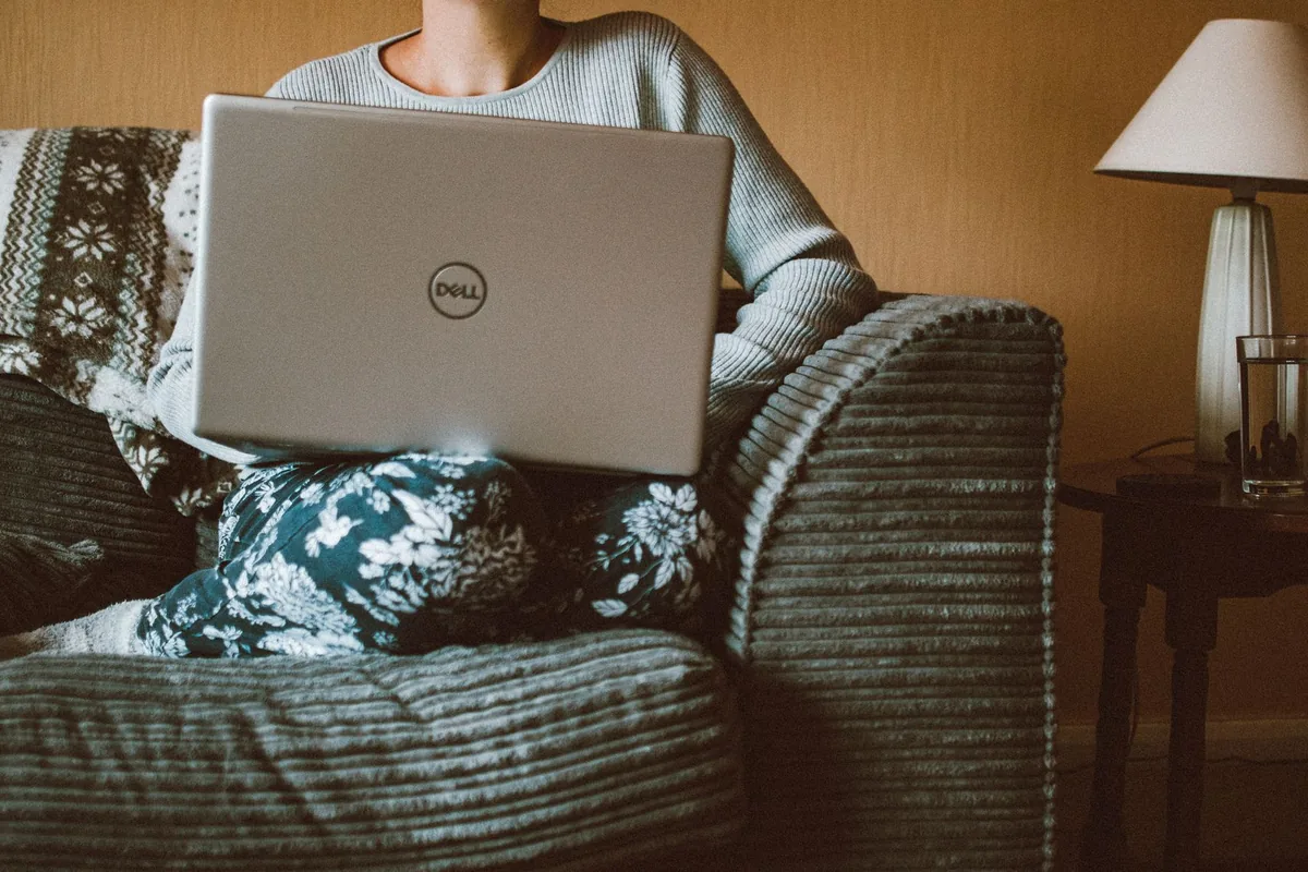 A woman in casual clothing works on a laptop while relaxing on a cozy sofa indoors.