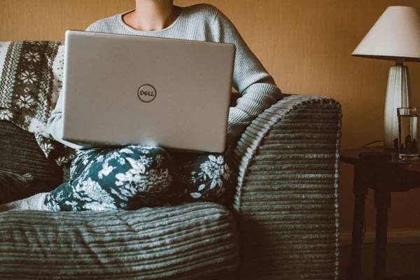 A woman in casual clothing works on a laptop while relaxing on a cozy sofa indoors.