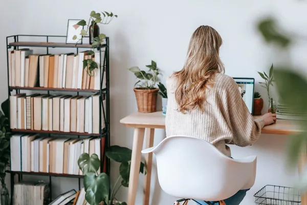 Back view of anonymous female remote worker sitting in armchair while browsing internet on netbook against bookshelves in house on light background