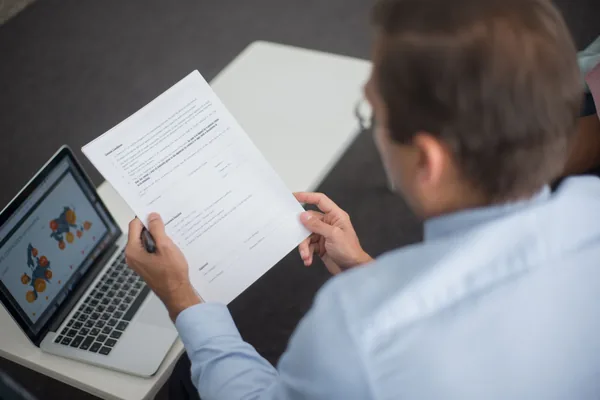 Professional in blue shirt reviewing documents next to laptop displaying charts.