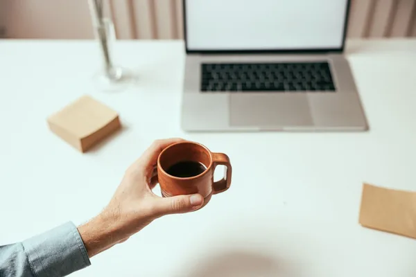 A person holds a brown coffee cup near a laptop on a clean white desk, perfect for remote work themes.