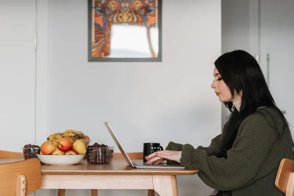 Side view of young female remote employee working on netbook at table with assorted fruits and hot drink at home