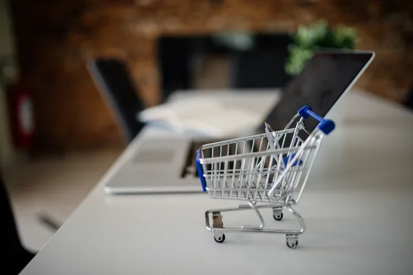 A miniature shopping cart on a table next to a laptop, suggesting online selling