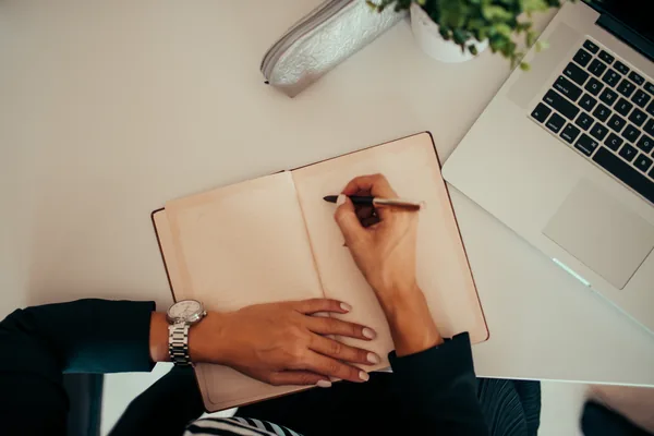 A person writing in a notebook beside a laptop at a modern workspace