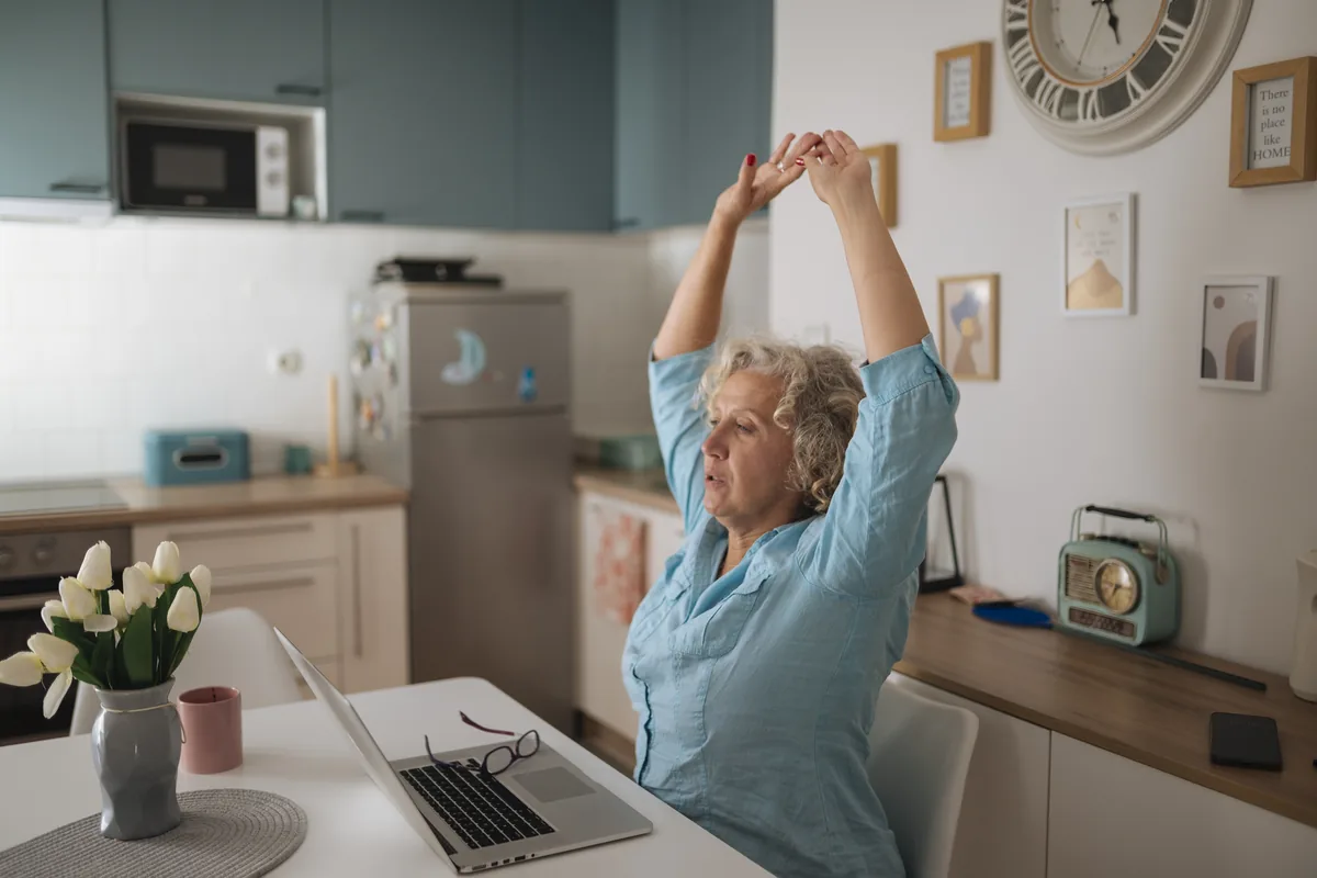 A person stretching at a home-office desk while working on a laptop